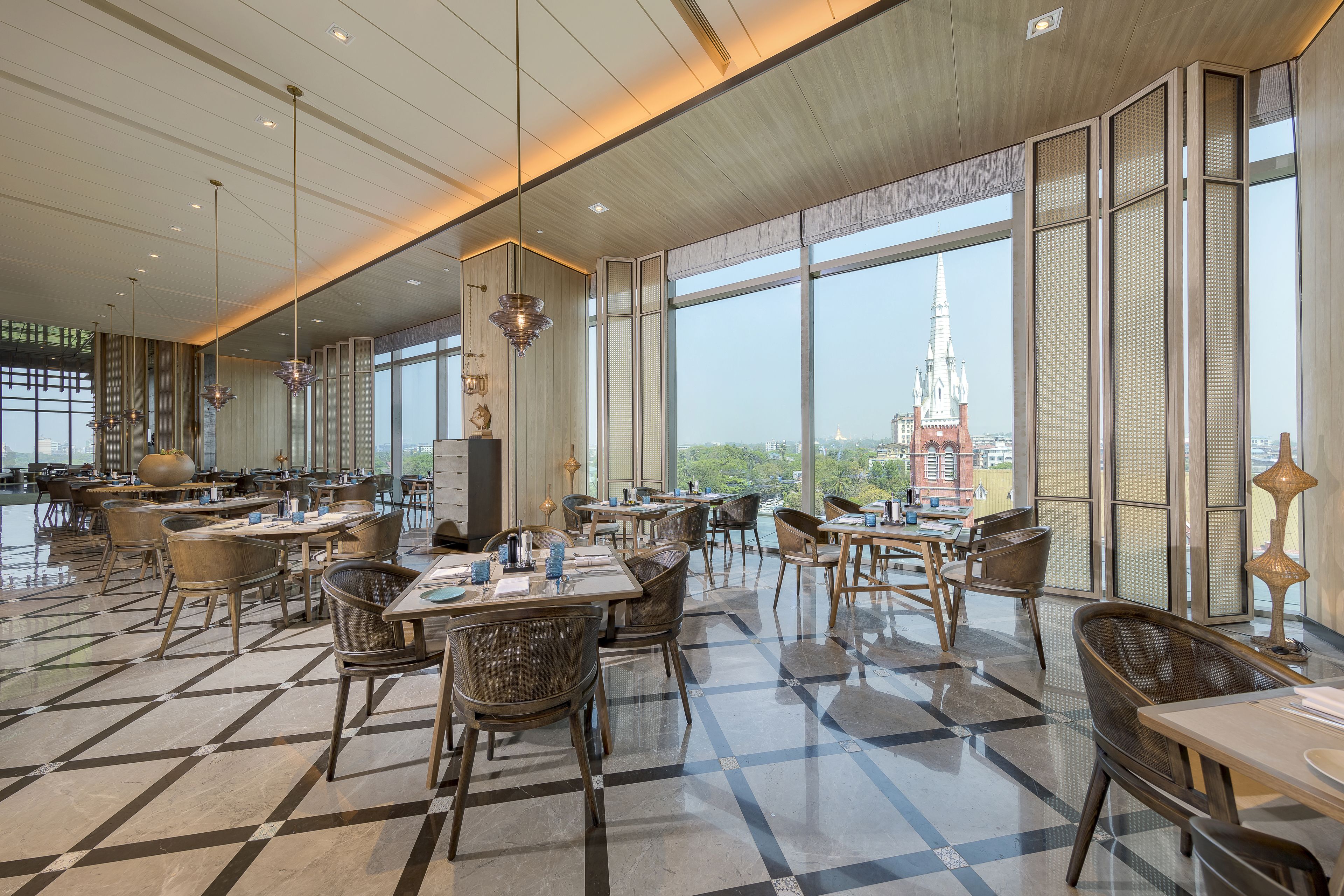 A spacious dining area with high ceilings, large windows, and a view of a distant church spire. Tables and chairs are arranged for dining.