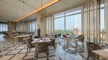 A spacious dining area with high ceilings, large windows, and a view of a distant church spire. Tables and chairs are arranged for dining.