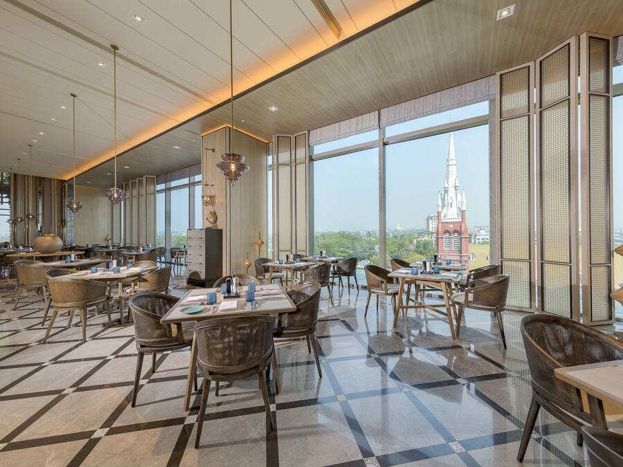 A spacious dining area with high ceilings, large windows, and a view of a distant church spire. Tables and chairs are arranged for dining.