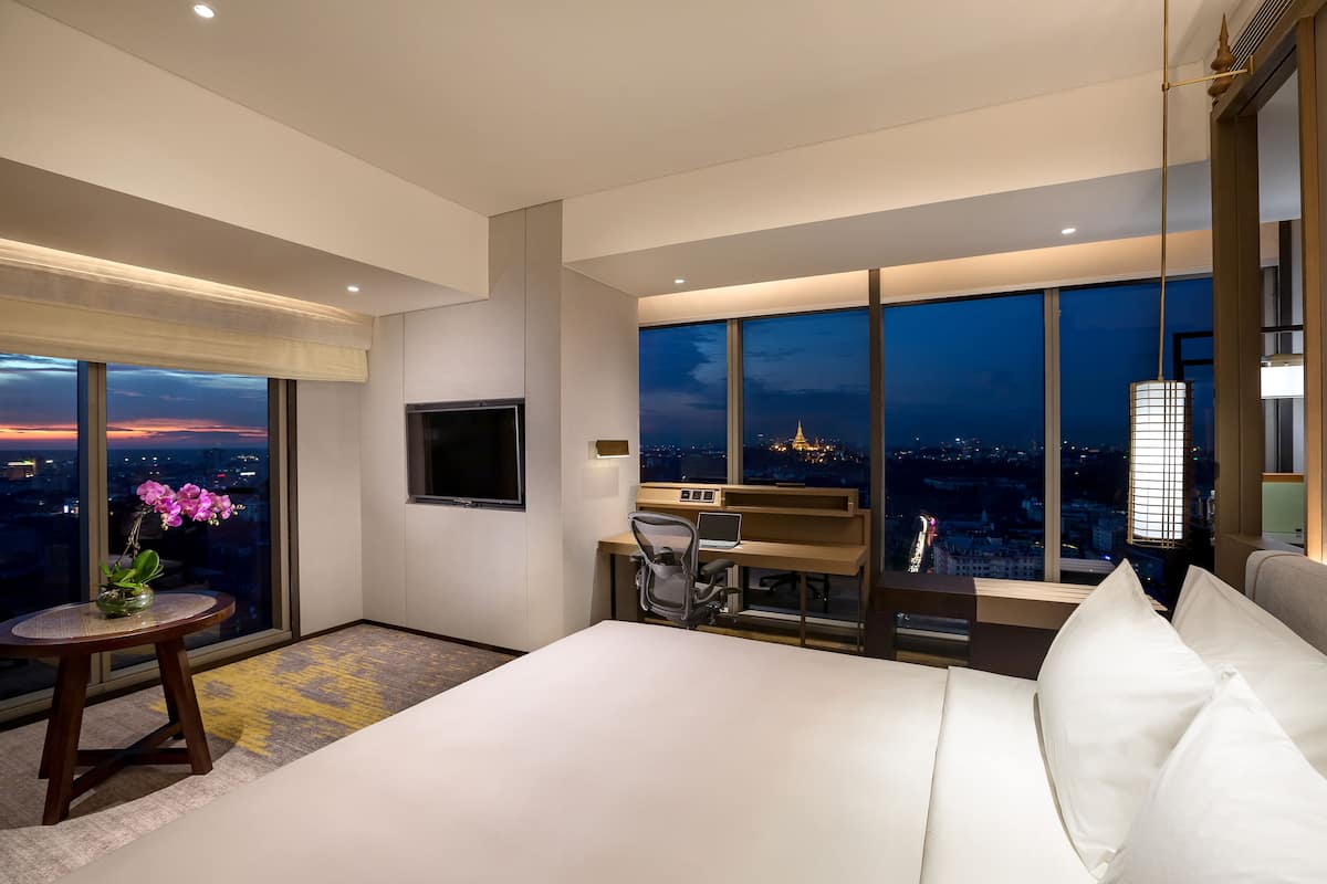A modern hotel room with a large bed, a desk, and a view of the city at night.