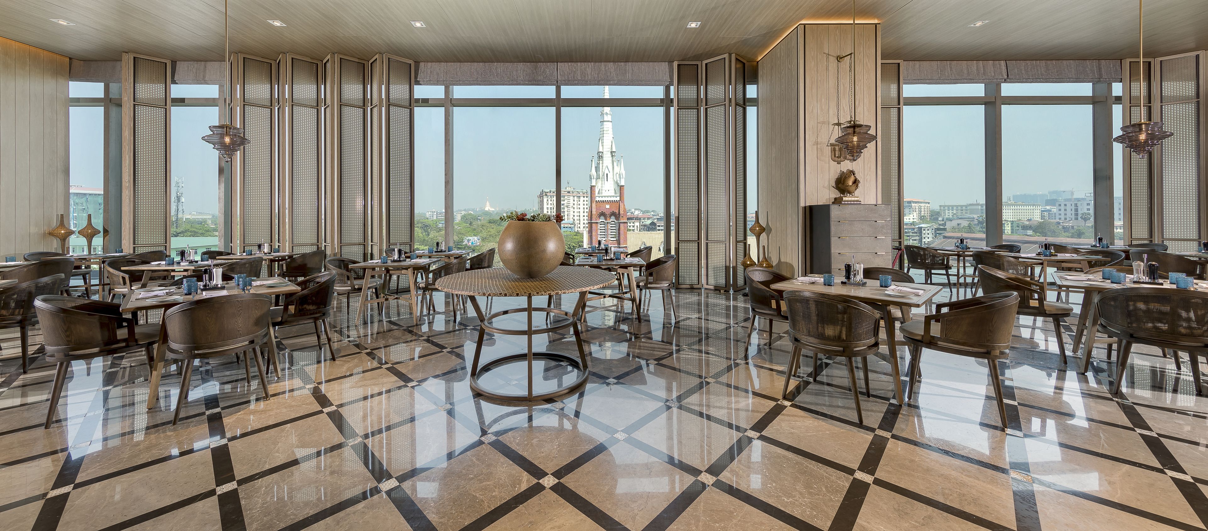A spacious dining area with a checkered floor, large windows offering a city view, and modern furnishings.