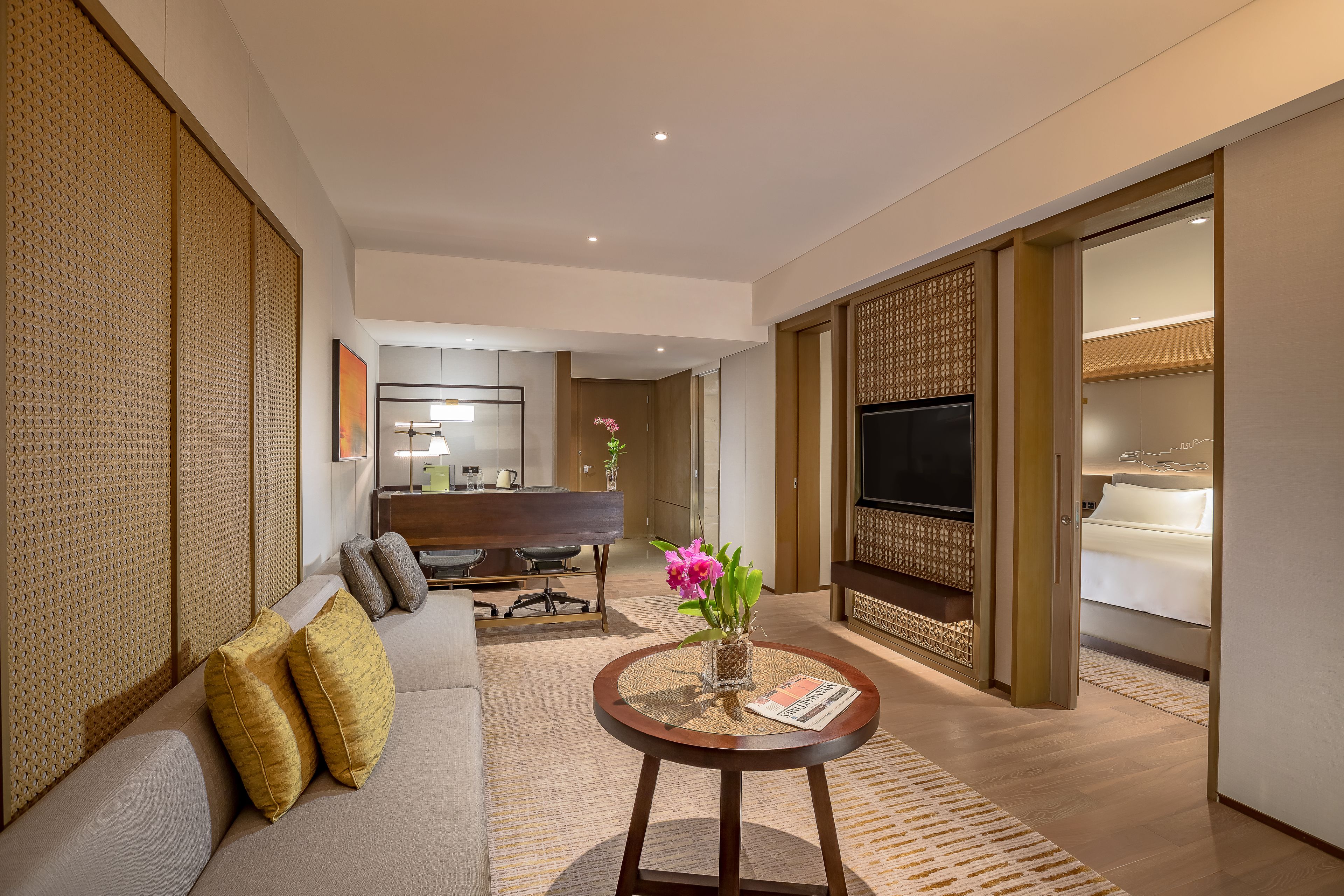 A modern hotel room with a sofa, a round wooden table with a vase and magazine, a TV, and a bed.