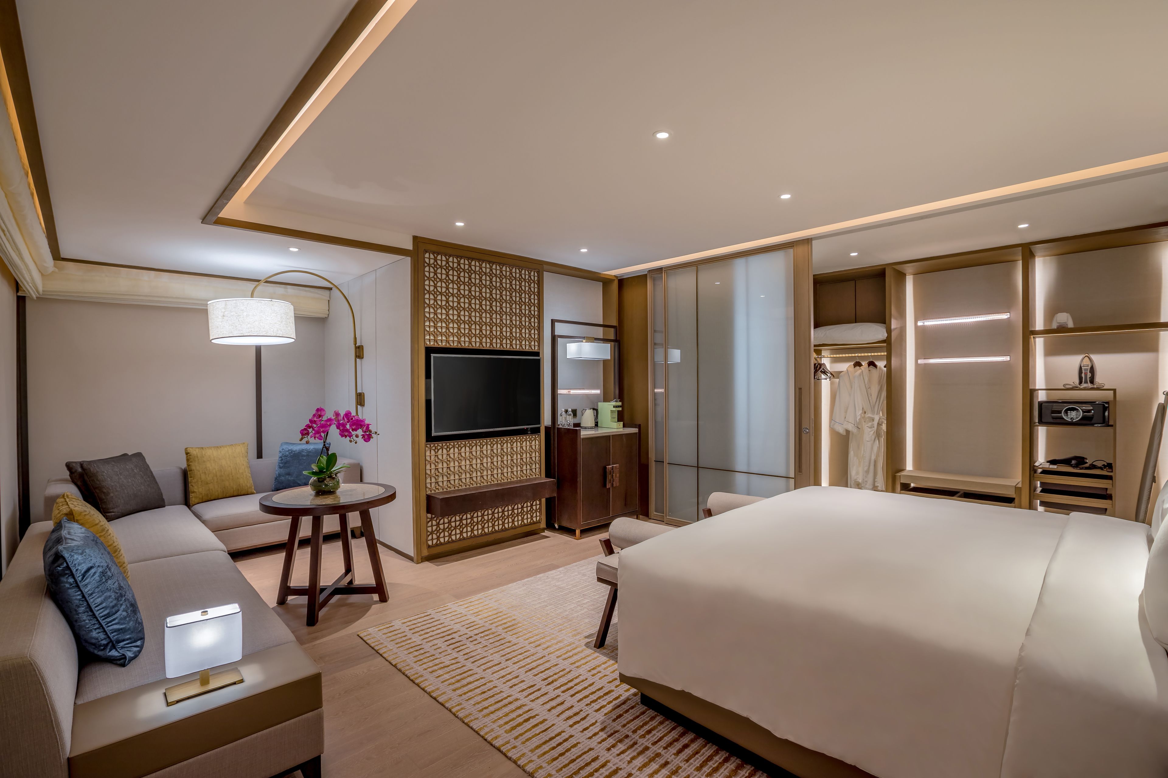 A modern hotel room with a large bed, a sofa, a TV, a wardrobe, and a small table with a lamp.