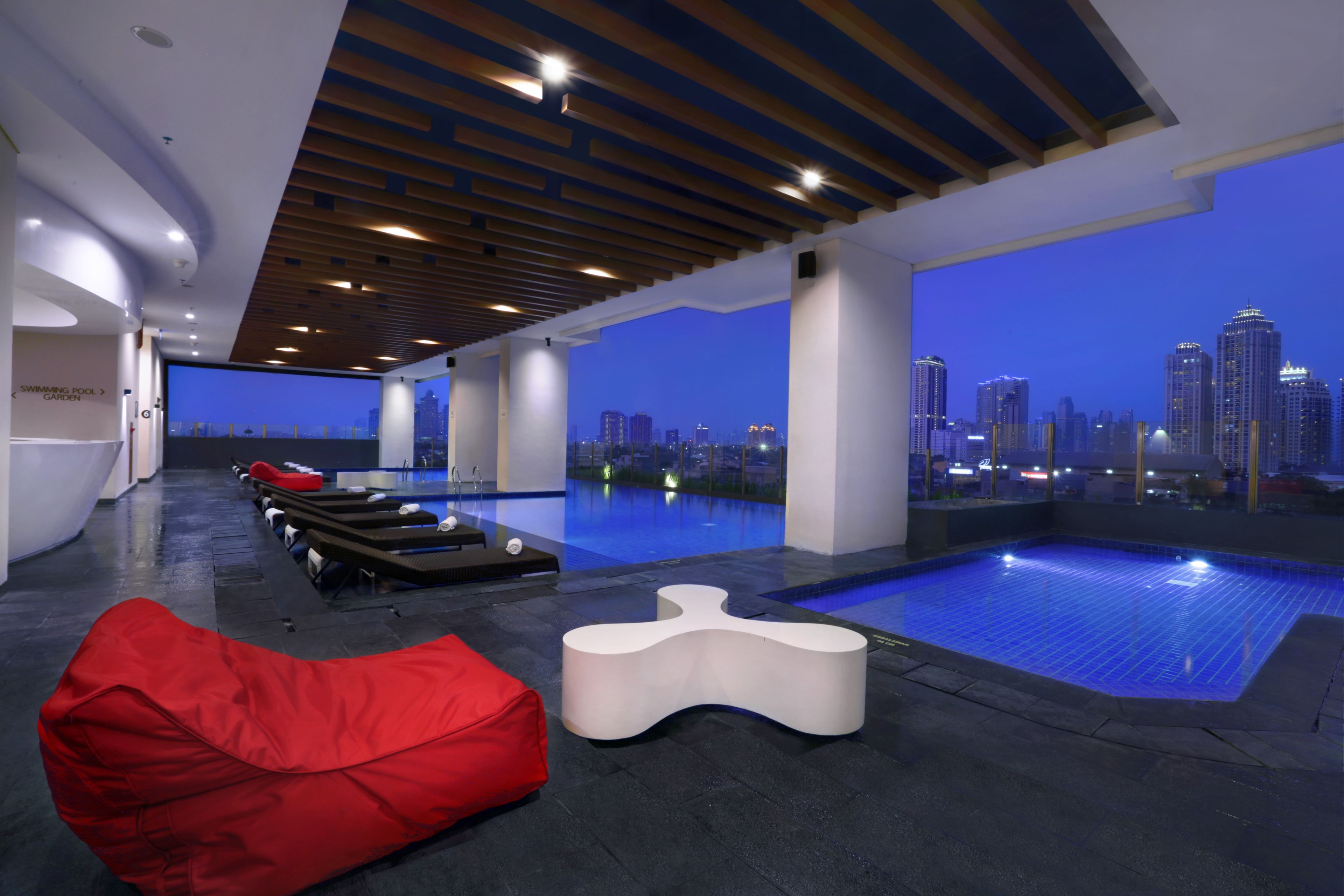 A modern indoor pool area with a city view, a red bean bag, and a white lounge chair.