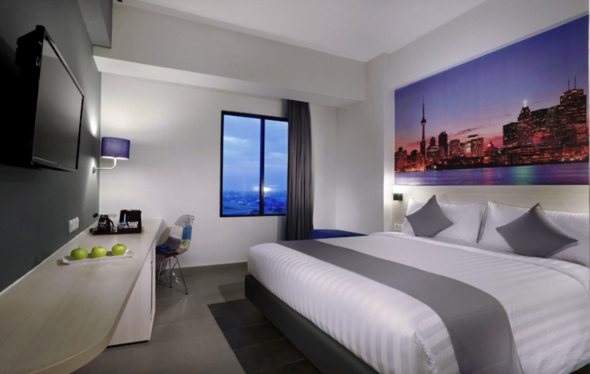 A hotel room with a bed, a desk, a chair, a window, and a cityscape painting.
