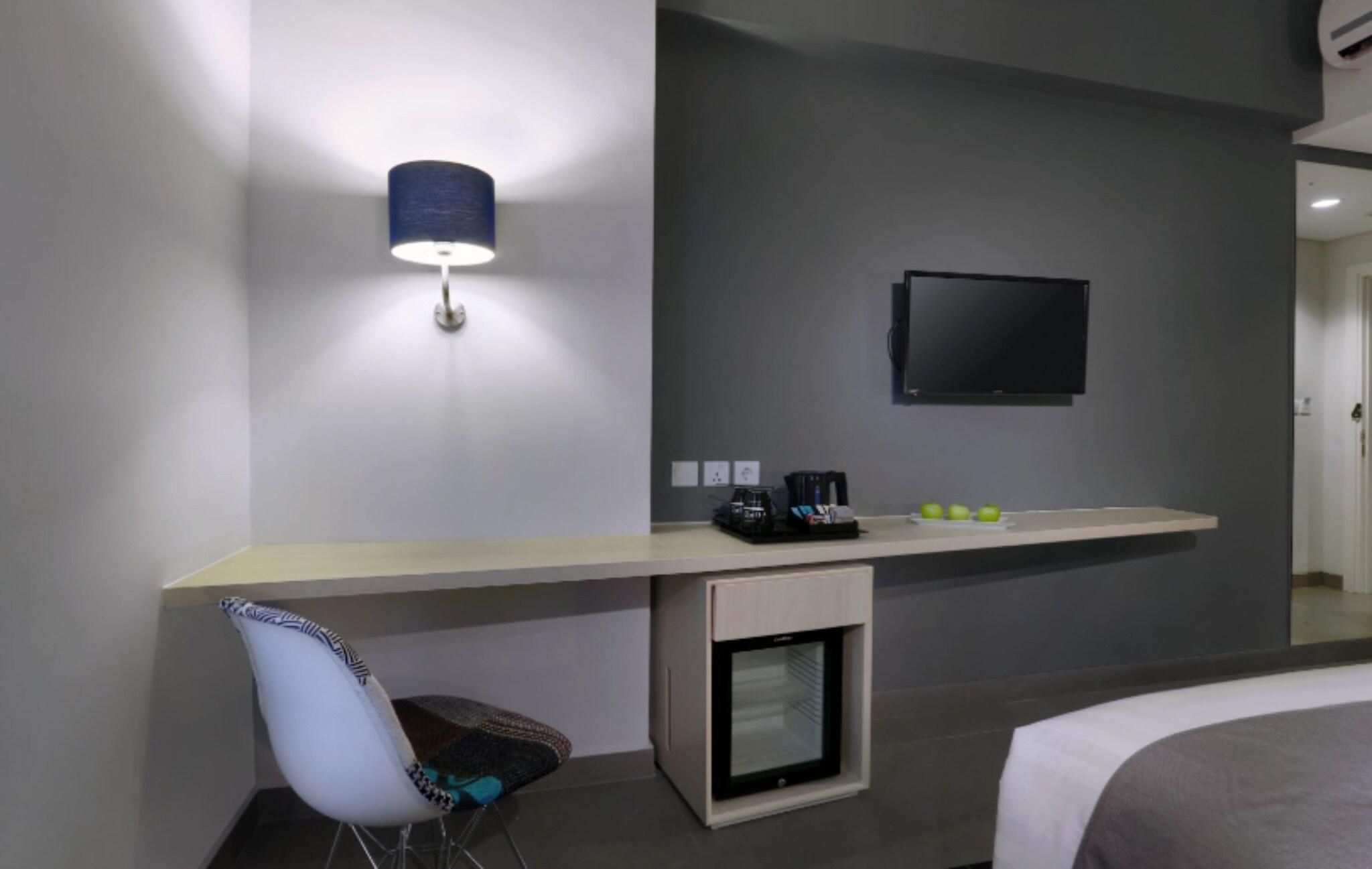 A modern hotel room with a flat-screen TV mounted on the wall, a small refrigerator, and a desk area with a chair.