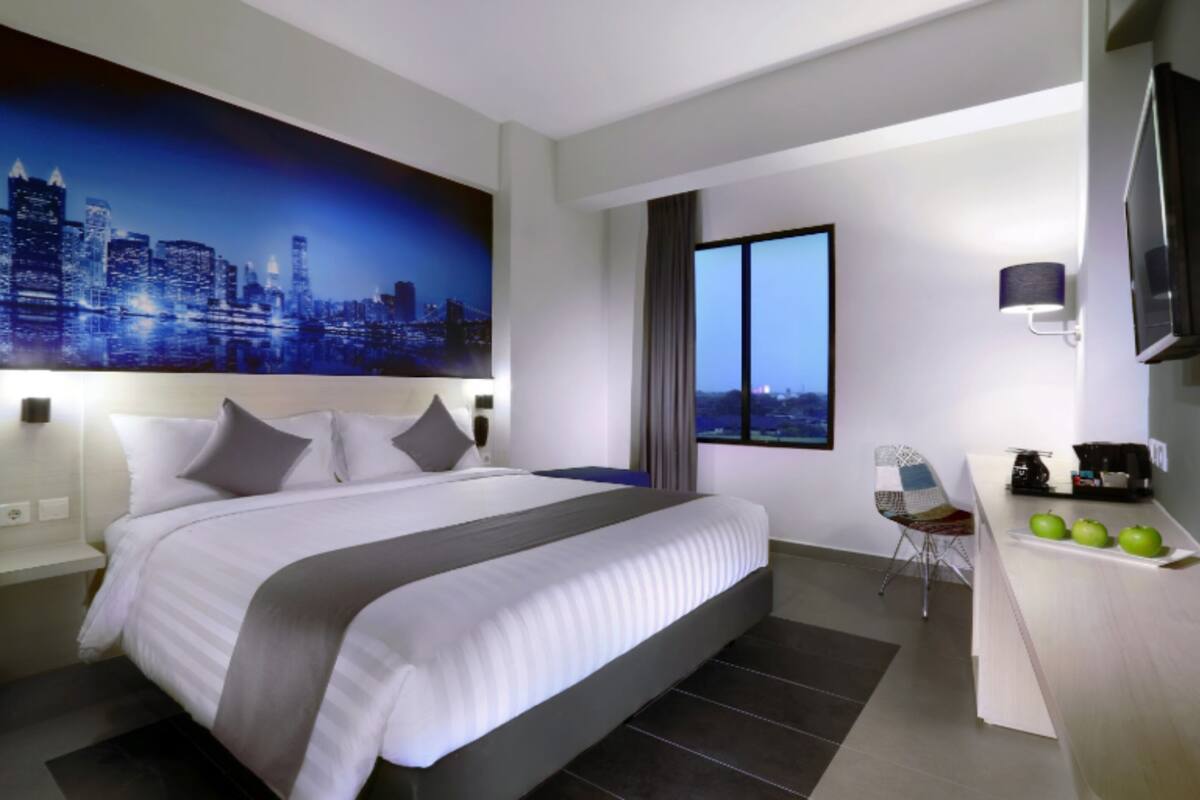 Ein modernes Hotelzimmer mit einem großen Bett, einem Stadtbild als Gemälde, einem Schreibtisch mit Lampe und Blick auf die Stadt durch das Fenster.