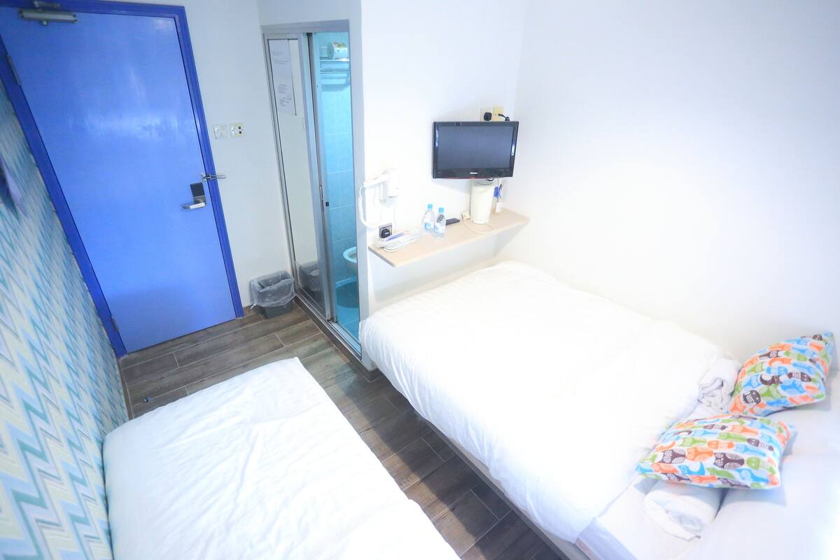A hotel room with a bed, a TV, a small table with a coffee maker, and a bathroom visible through a glass door.