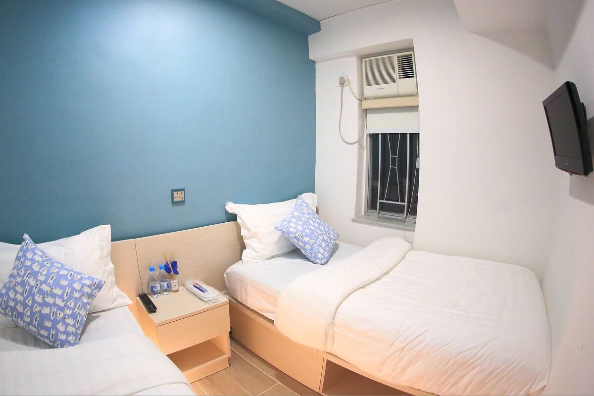A hotel room with two beds, a TV mounted on the wall, an air conditioning unit, and a window with blinds.