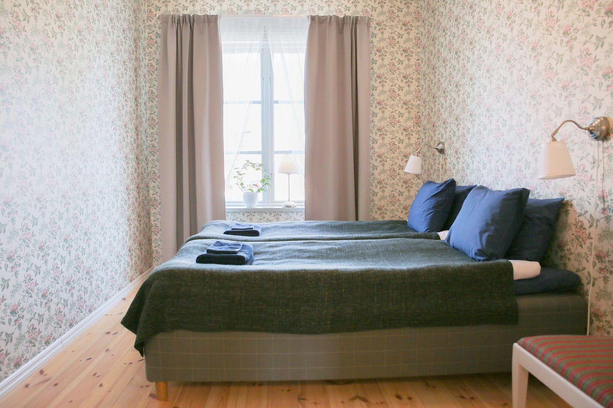 Une pièce avec un papier peint à motifs floraux, un lit avec une literie bleue, une lampe de chevet et une fenêtre avec des rideaux.