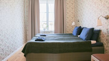 A room with floral wallpaper, a bed with blue bedding, a bedside lamp, and a window with curtains.