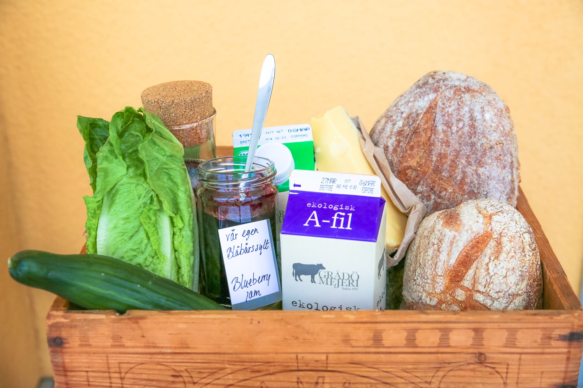 A wooden crate containing a variety of food items including bread, cheese, a jar of jam, and leafy greens.