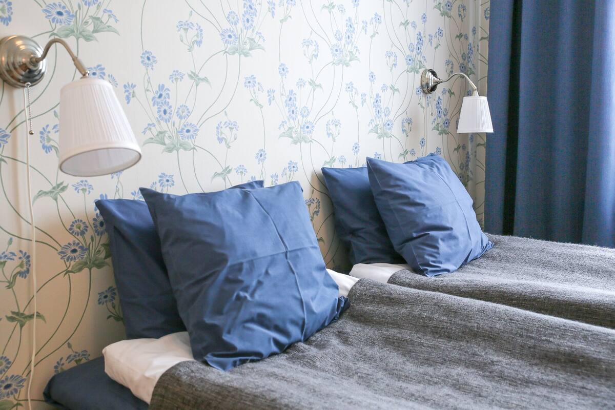 Un lit avec des oreillers bleus et une couverture grise, un mur recouvert d’un papier peint à motifs floraux et deux lampes fixées au mur.