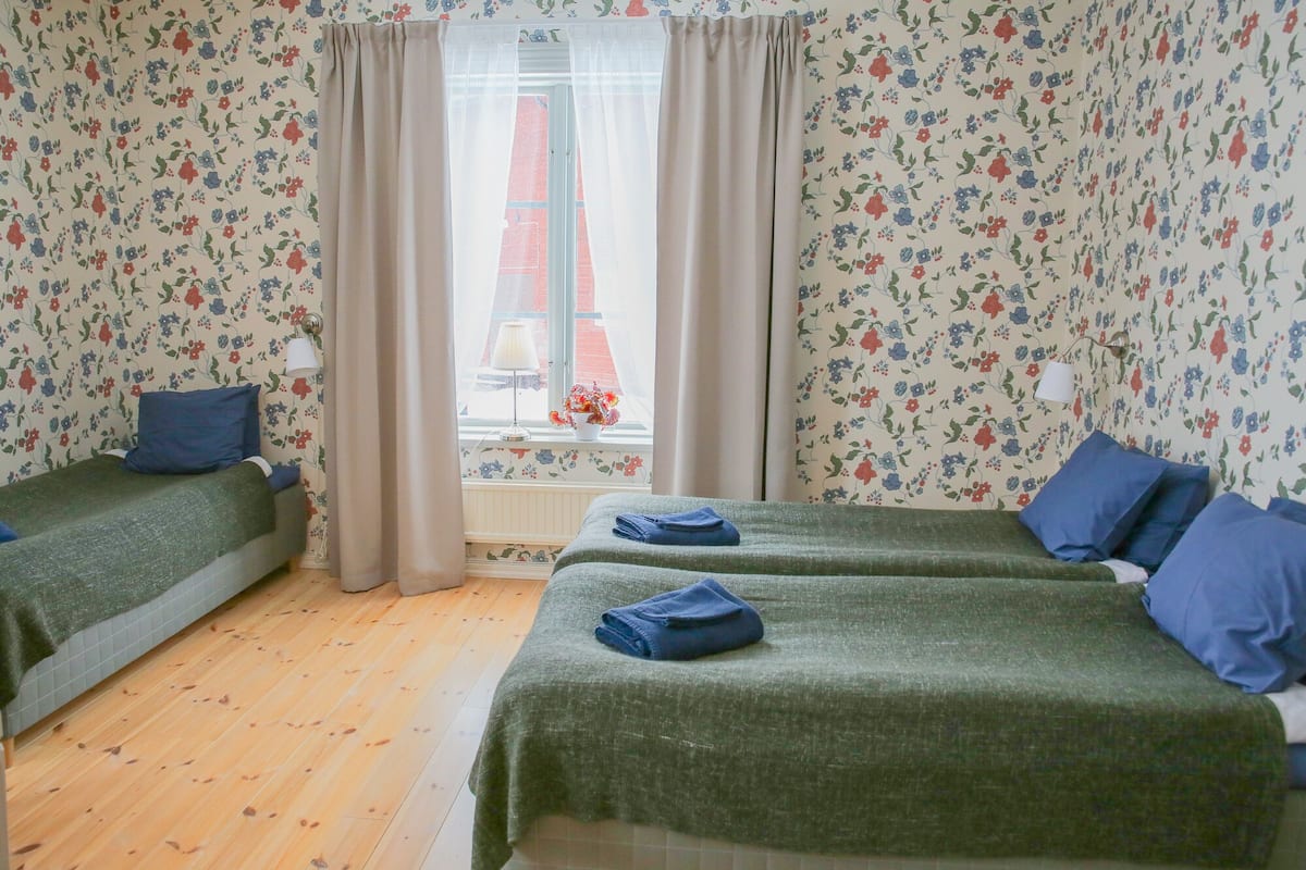 Une chambre avec deux lits, un papier peint à motifs floraux, une fenêtre avec des rideaux et une lampe.