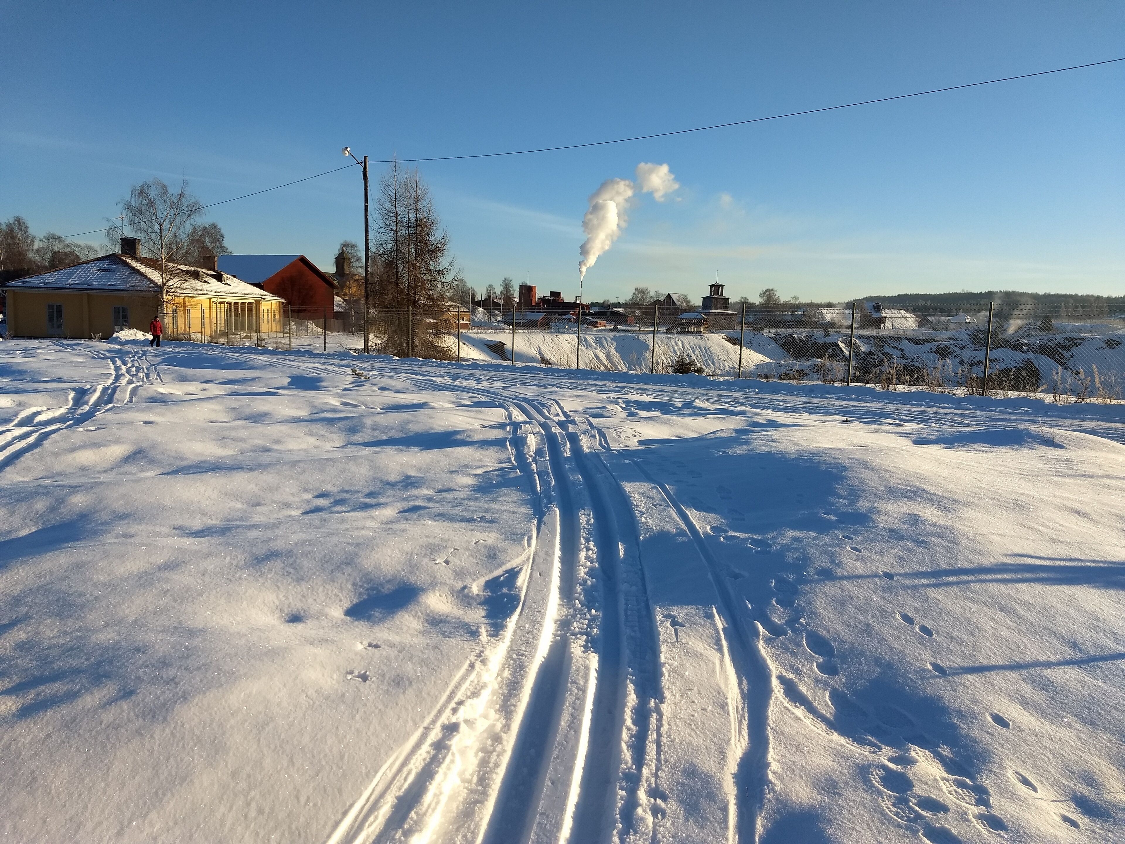 A snowy landscape with a clear sky, a building with a yellow facade, and a chimney emitting smoke.