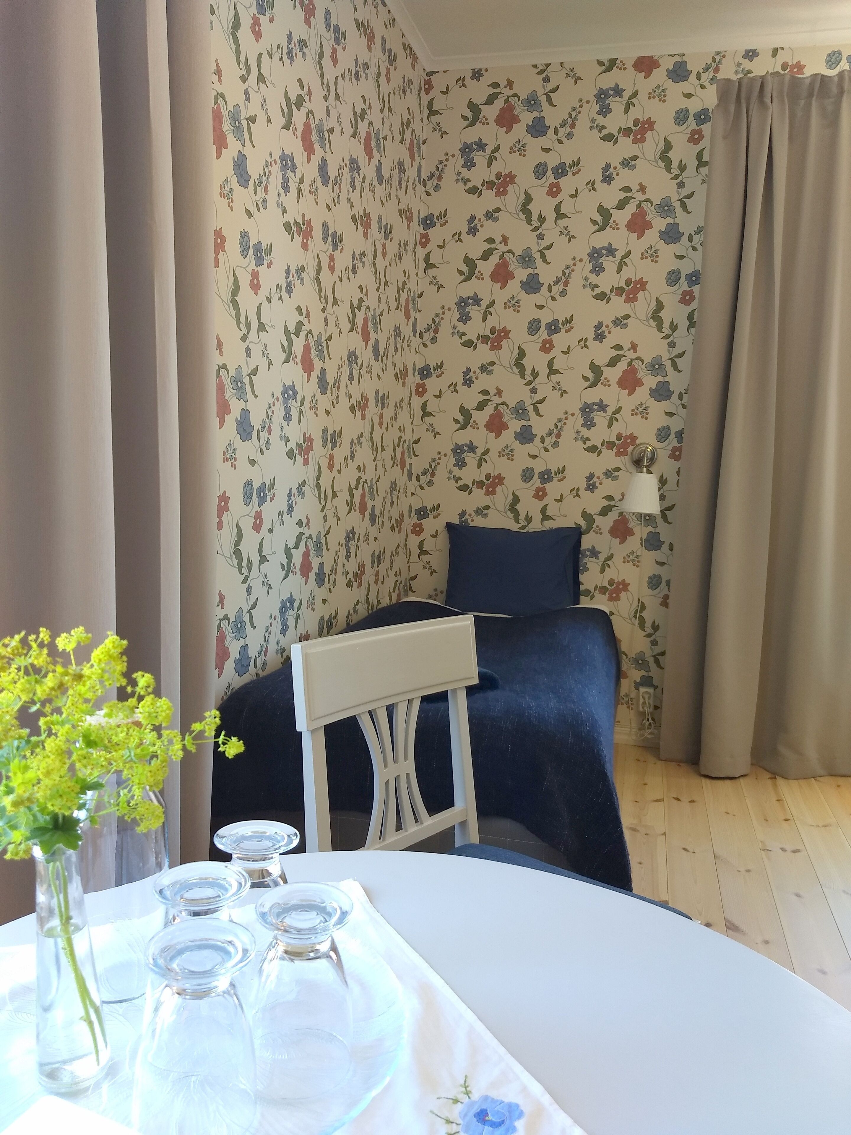 A hotel room with floral wallpaper, a bed with a dark blue cover, a white chair, a table with a vase of flowers, and a lamp.
