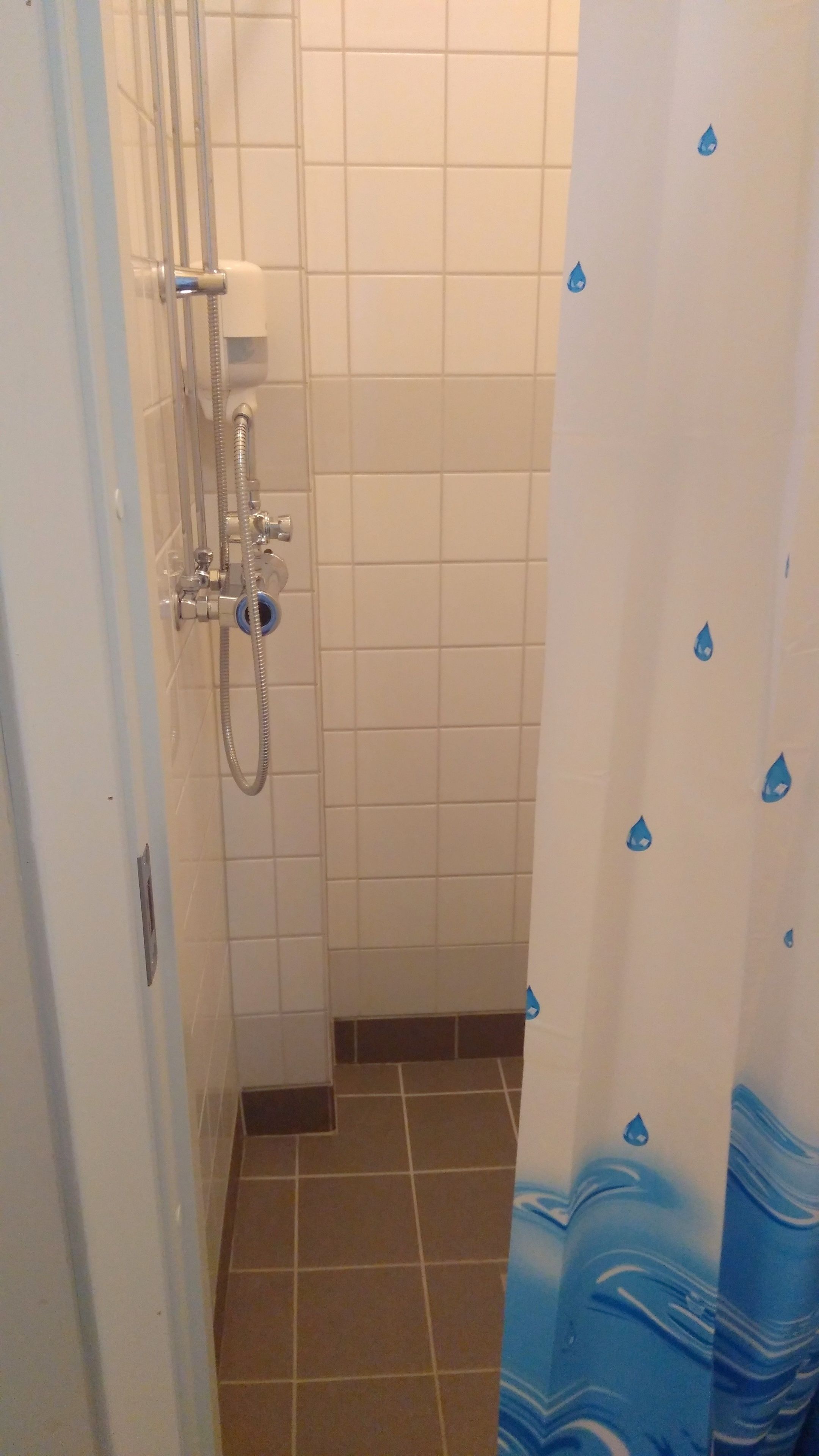 Un baño pequeño con azulejos blancos, una ducha con cabezal de ducha de mano y una cortina de ducha azul con diseños de gotas de agua.