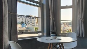 Ein Zimmer mit zwei weißen Stühlen und einem kleinen runden Tisch, großen Fenstern mit Vorhängen und Blick auf Gebäude.