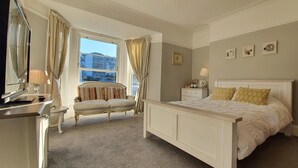 Ein Schlafzimmer mit einem großen Bett, einem Nachttisch, einem Sessel, einer Kommode und einem Fenster mit Vorhängen.