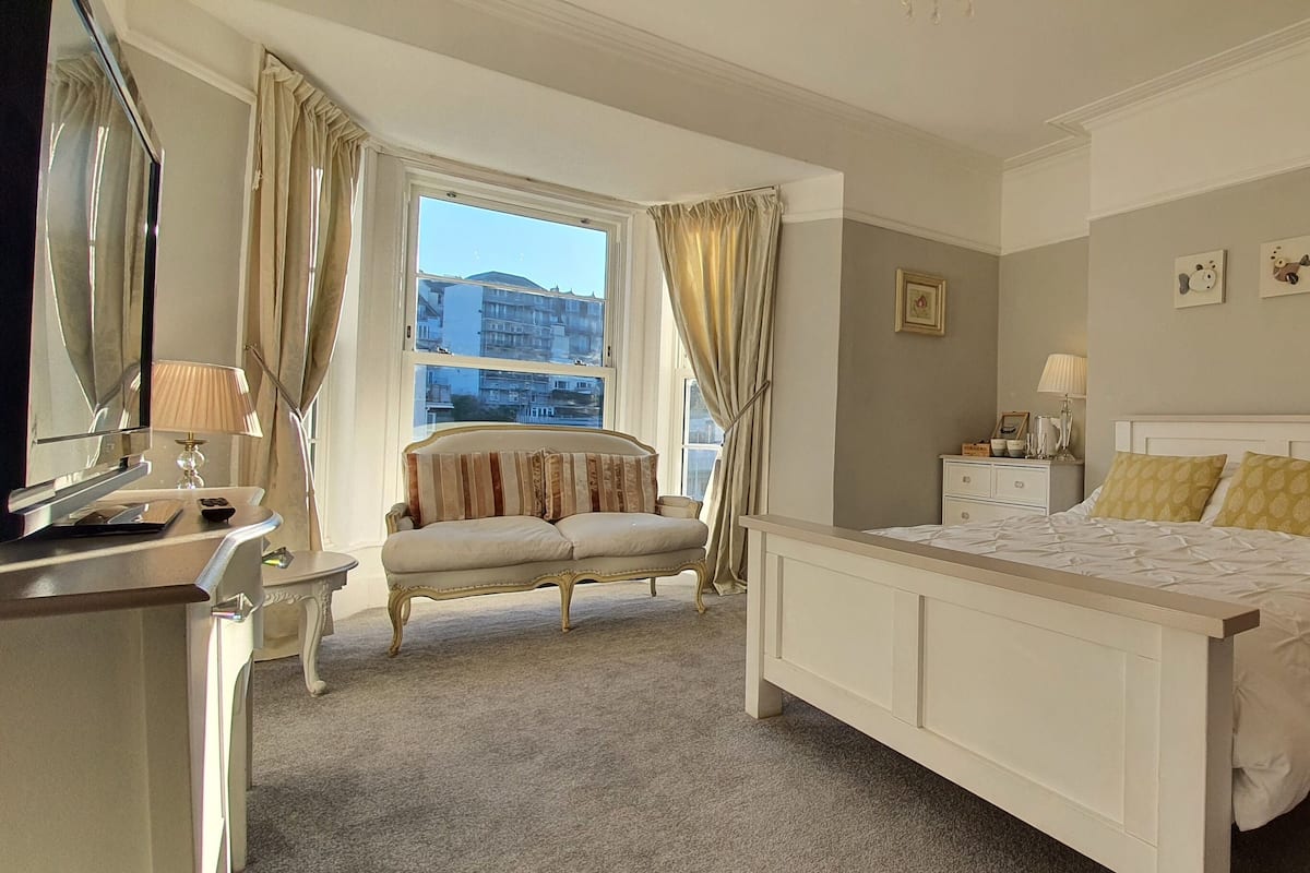 Une chambre à coucher avec un grand lit, une table de chevet, une chaise, une commode et une fenêtre avec des rideaux.