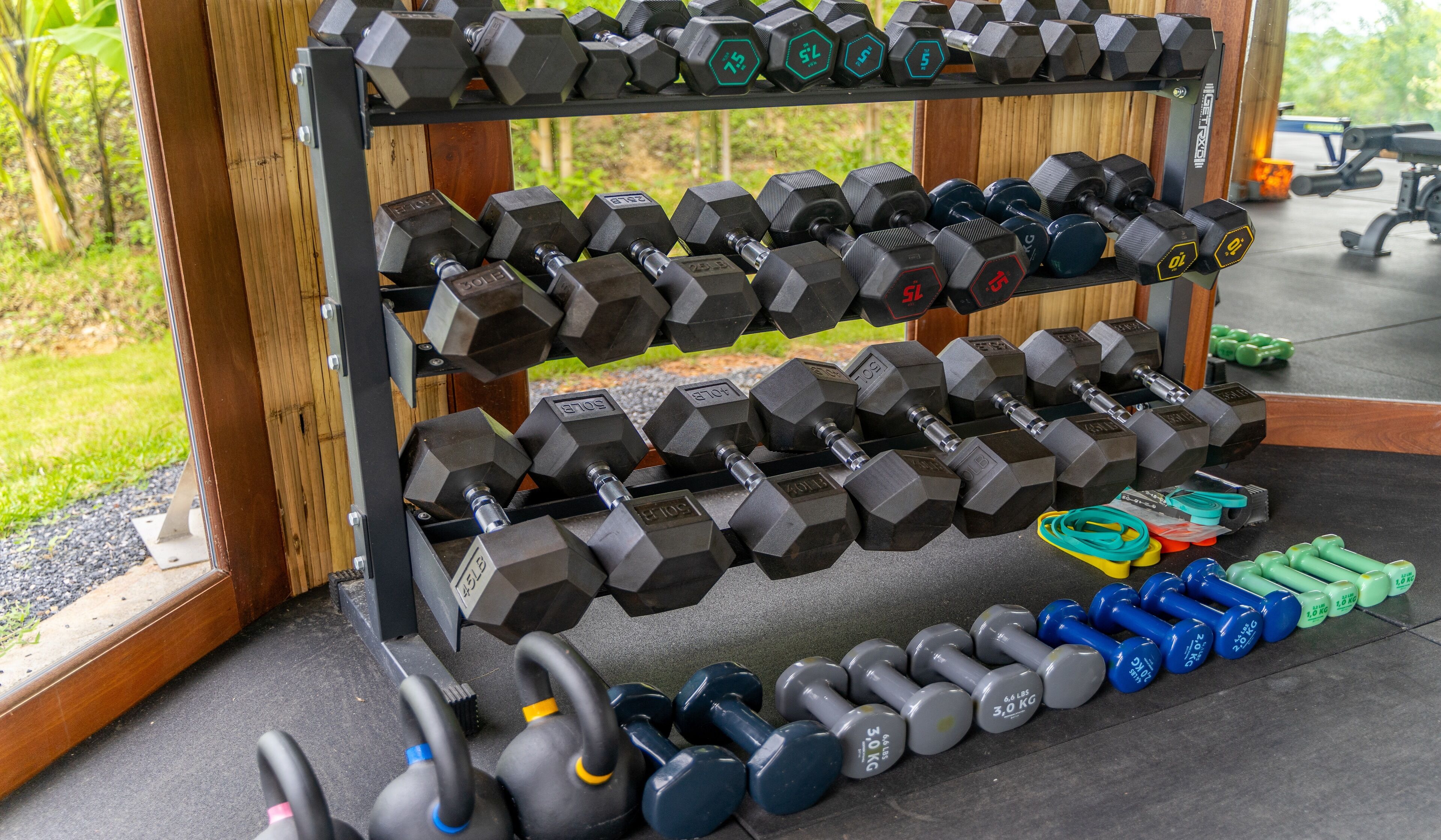 A rack of hexagonal dumbbells with weights ranging from 5 to 15 pounds, arranged on a wooden floor with a view of greenery outside.