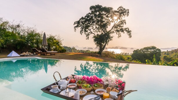 Desayuno servido en una mesa junto a la piscina, con diversos alimentos y bebidas, rodeado de abundante vegetación y vistas al agua.