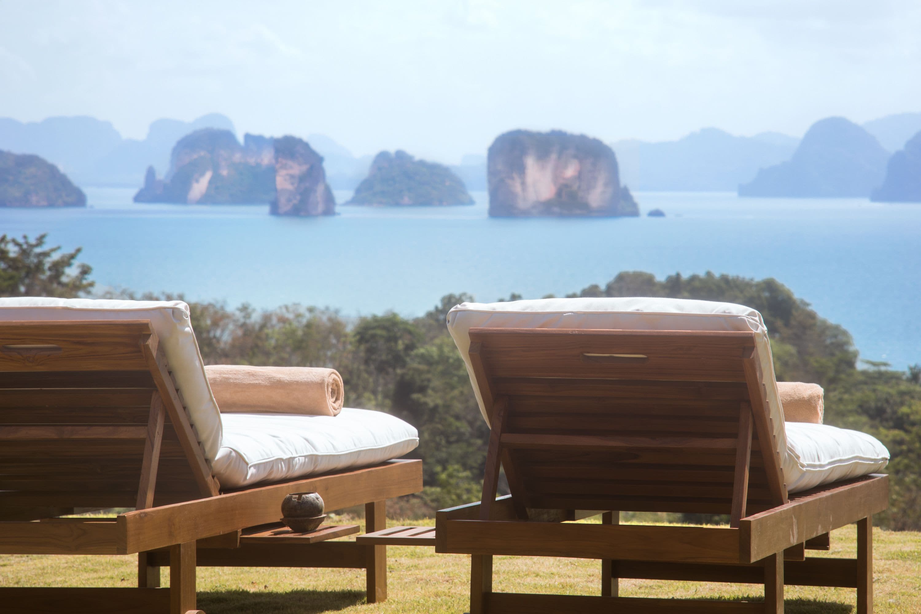 Dos sillones de madera con cojines, ubicados en un área verde con vistas al mar tranquilo y a islas distantes.