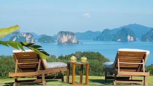 Dos sillones de madera con cojines, una mesita con dos bebidas verdes, y vistas a un paisaje costero con islas y agua azul cristalina.