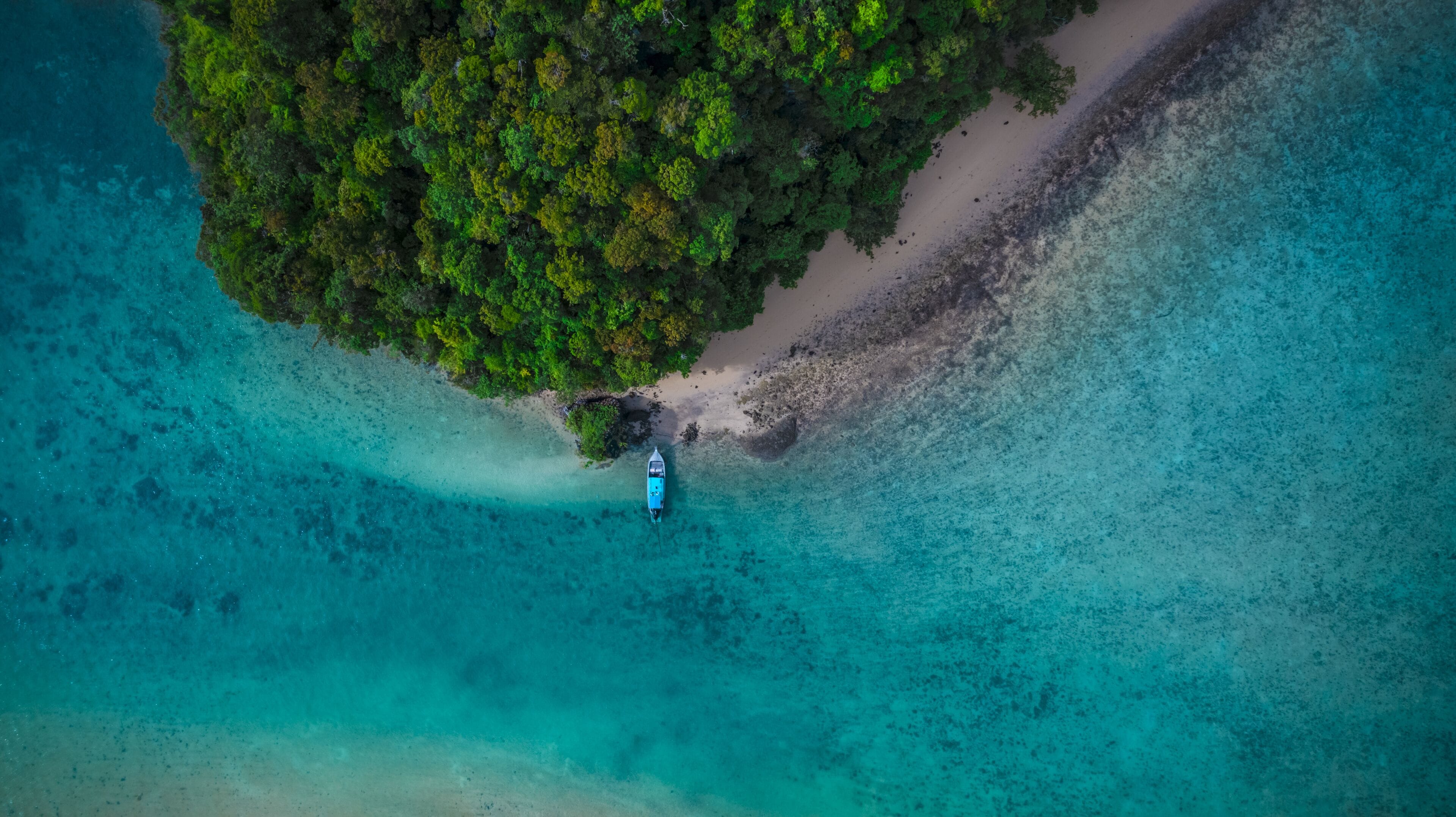 An aerial view of a tropical island with a boat anchored near the shore, surrounded by clear turquoise waters and dense green foliage.