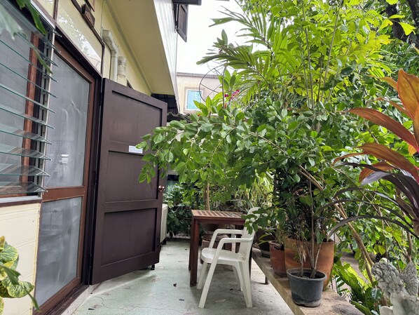 Un pequeño espacio al aire libre con una mesa y sillas, rodeado de plantas en macetas y una puerta marrón grande.