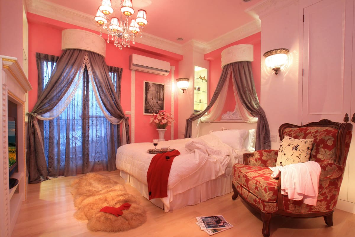 A bedroom with a large bed, a chair, a window with curtains, a chandelier, and a painting on the wall.
