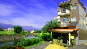 Un edificio residencial de varios pisos con un balcón en el segundo piso, una pequeña zona de estar al aire libre con plantas, y vistas a un río y montañas al fondo.
