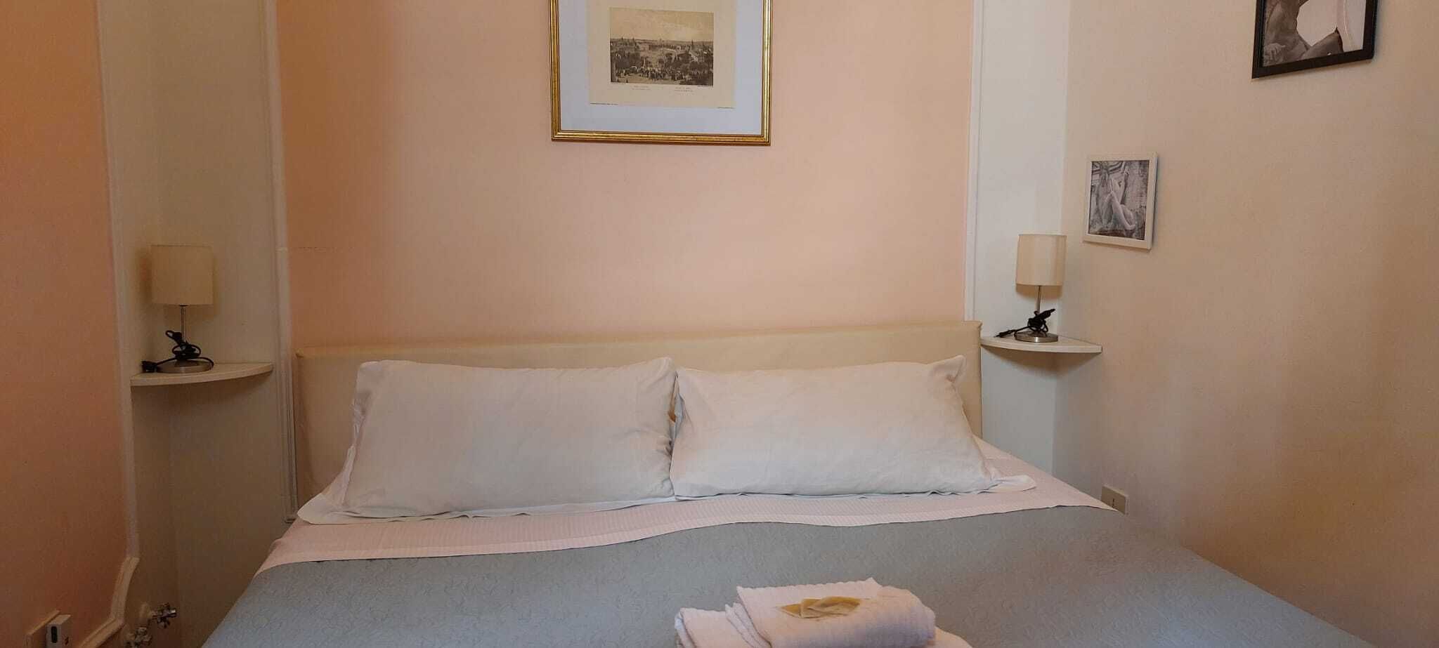 A neatly made bed with two pillows and a folded towel, a bedside lamp on each side, and a framed picture on the wall above the bed.