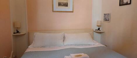 A neatly made bed with two pillows and a folded towel, a bedside lamp on each side, and a framed picture on the wall above the bed.
