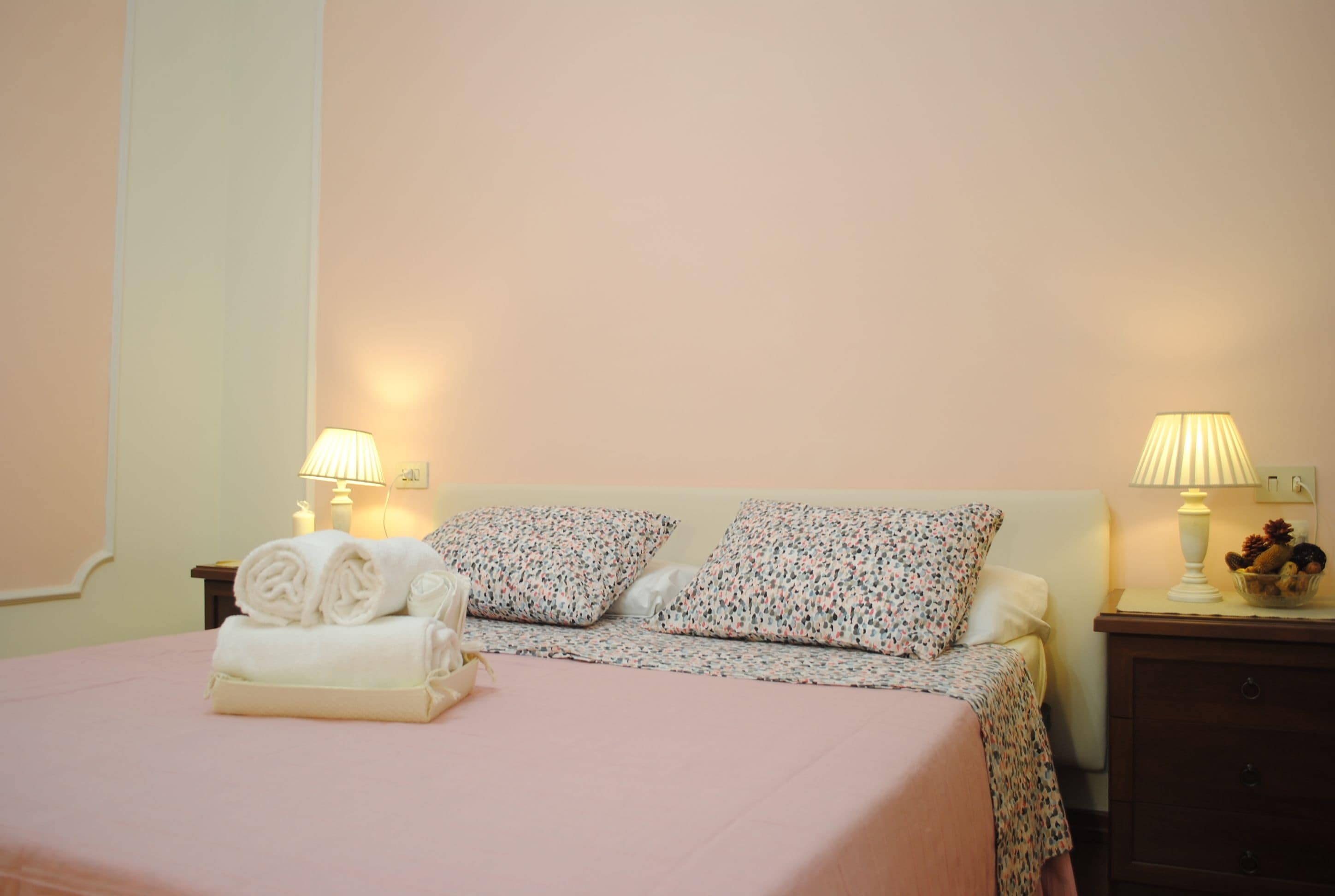 A neatly made bed with rolled towels, two floral-patterned pillows, and two bedside lamps on a nightstand.