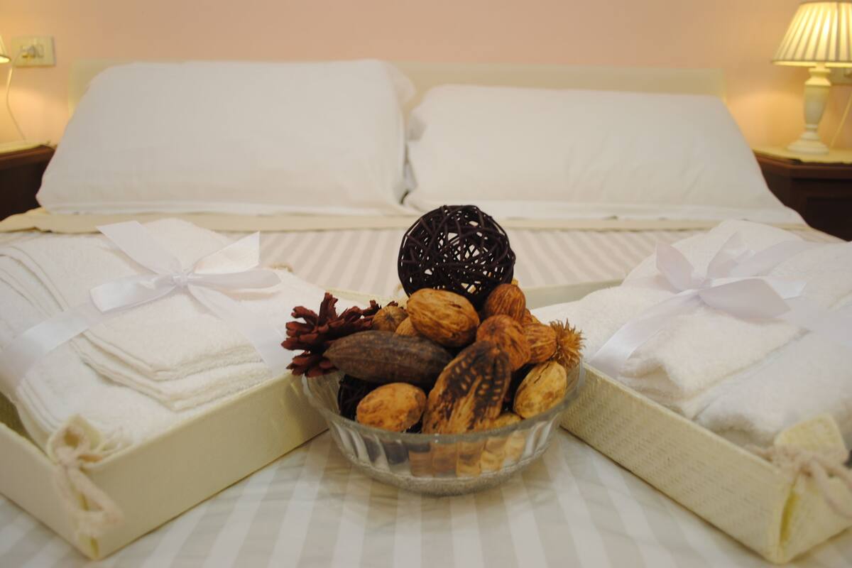 A neatly made bed with white linens and pillows, a glass bowl with nuts and dried flowers, and a bedside lamp on the nightstand.