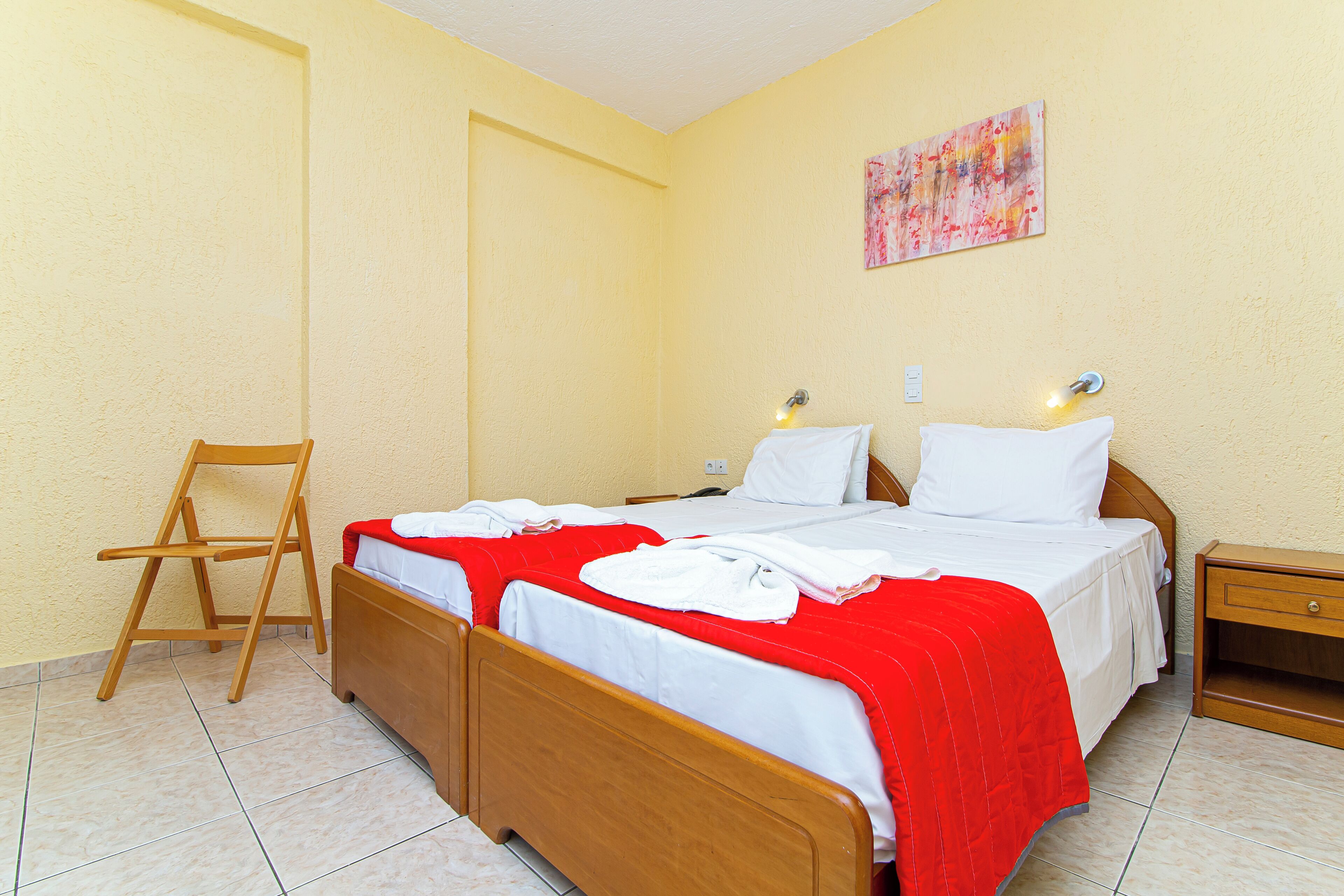 A hotel room with two wooden beds, a red and white bedspread, a wooden nightstand with a lamp, and a wooden chair.