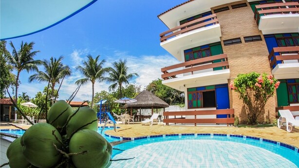Zona de piscina con ambiente tropical, que incluye un edificio con varios balcones y una estructura de techo de paja.
