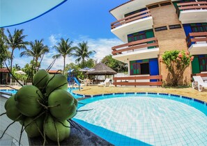 A pool area with a tropical setting, including a building with multiple balconies and a thatched-roof structure. - Hotel Rio Mar (Barra de São Miguel)