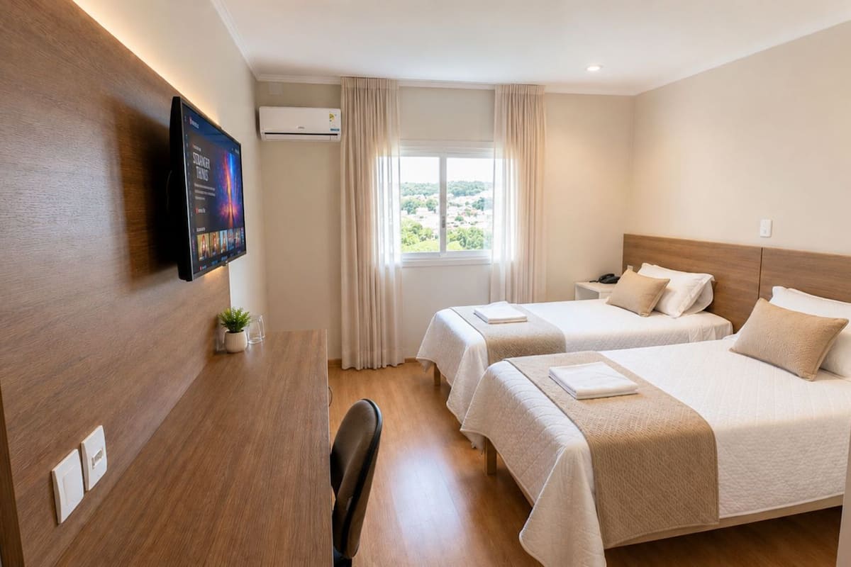 A hotel room with two beds, a desk with a TV, a window with curtains, and an air conditioning unit.