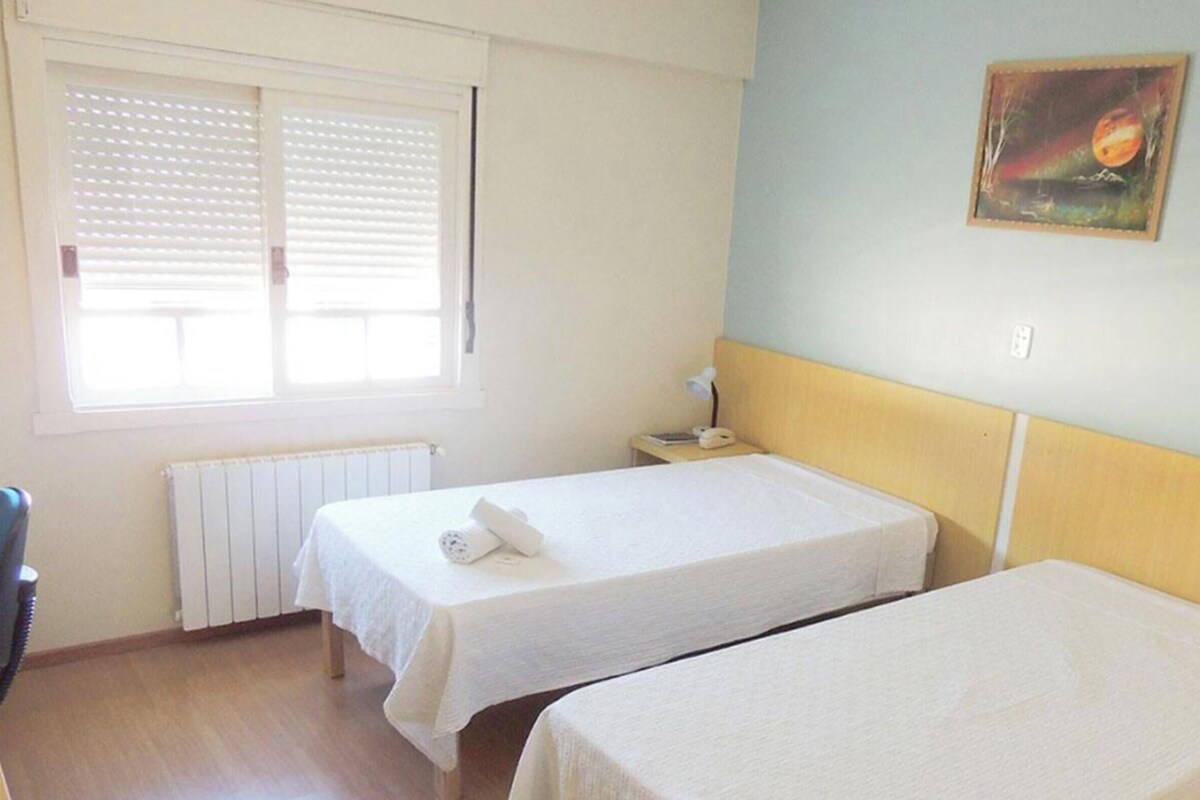 A room with two single beds, a radiator, a window with blinds, a desk with a lamp, and a painting on the wall.
