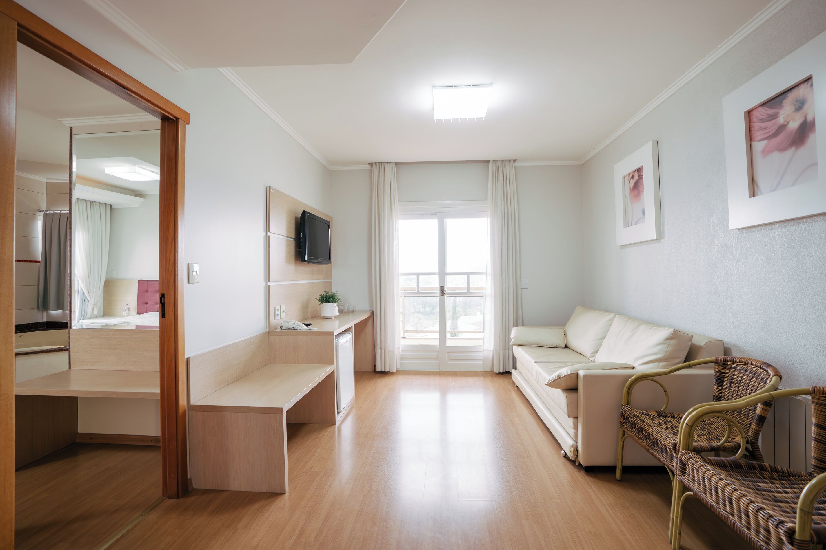 A modern room with a bed, a desk, a sofa, a wicker chair, a TV, and a window with curtains.