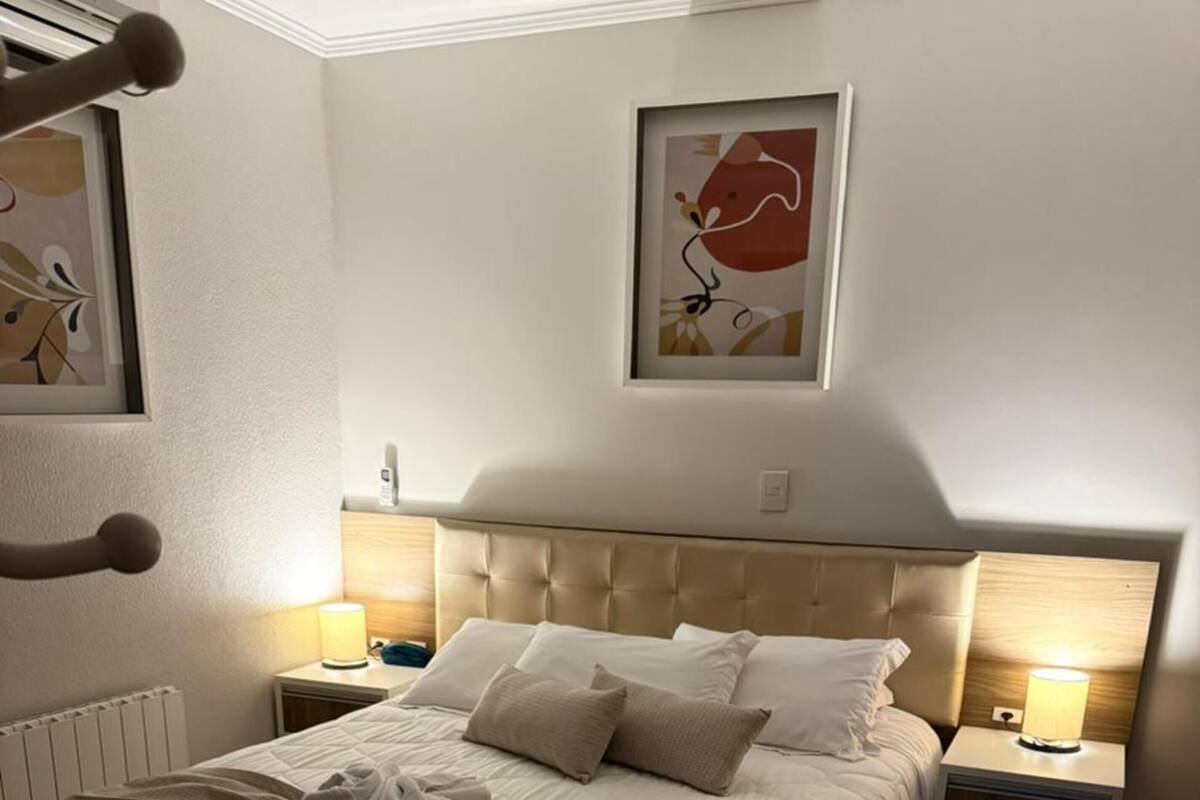 A neatly made bed with a quilted cover, two bedside lamps, a headboard, and a framed abstract painting on the wall.
