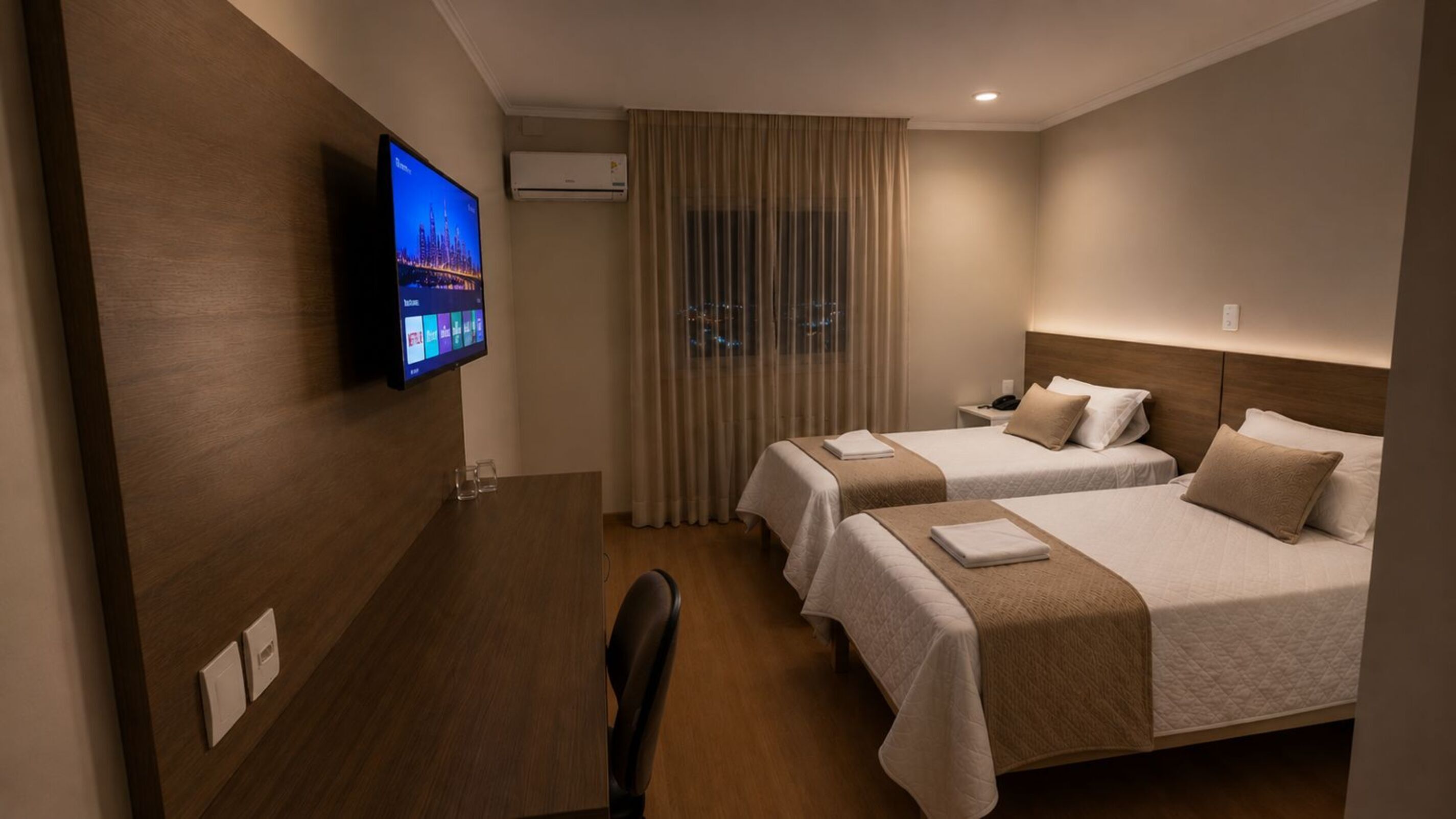 A hotel room with two beds, a desk, a TV, and an air conditioning unit.