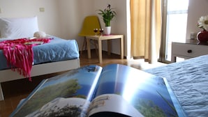 Une chambre d’hôtel avec deux lits, une petite table ornée d’une plante et de deux verres à vin, et une fenêtre avec des rideaux.
