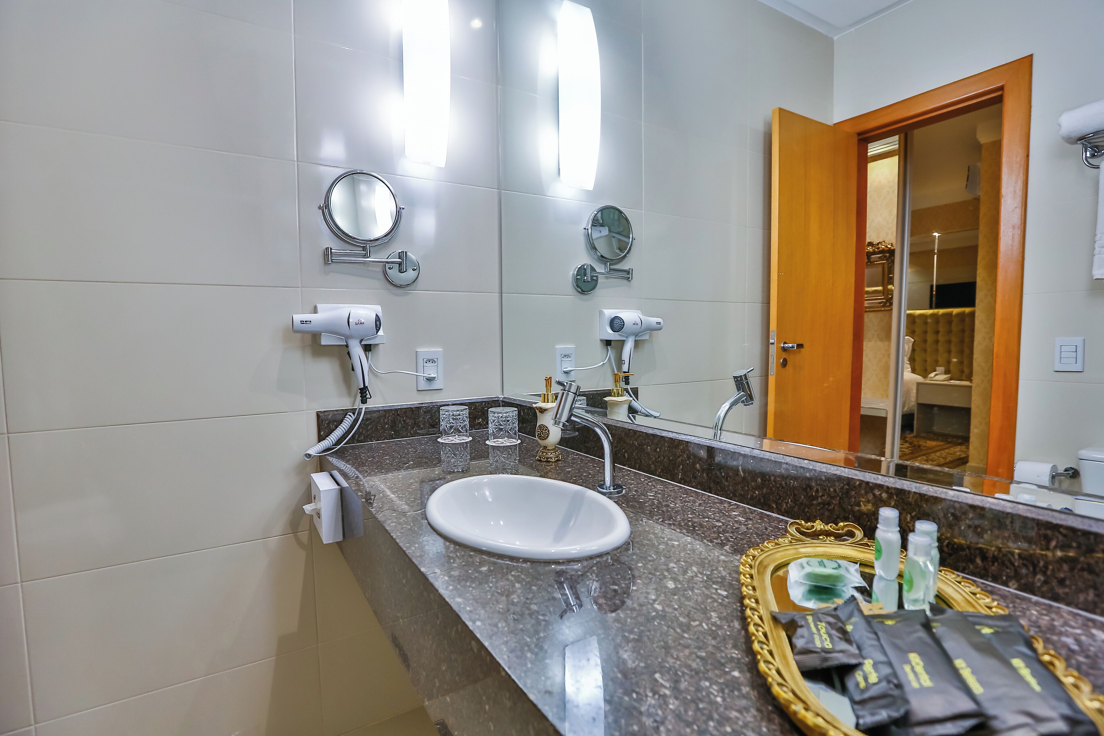 Casa de banho com uma pia branca, dois espelhos, um espelho sobre a penteadeira e uma penteadeira com produtos de higiene pessoal.