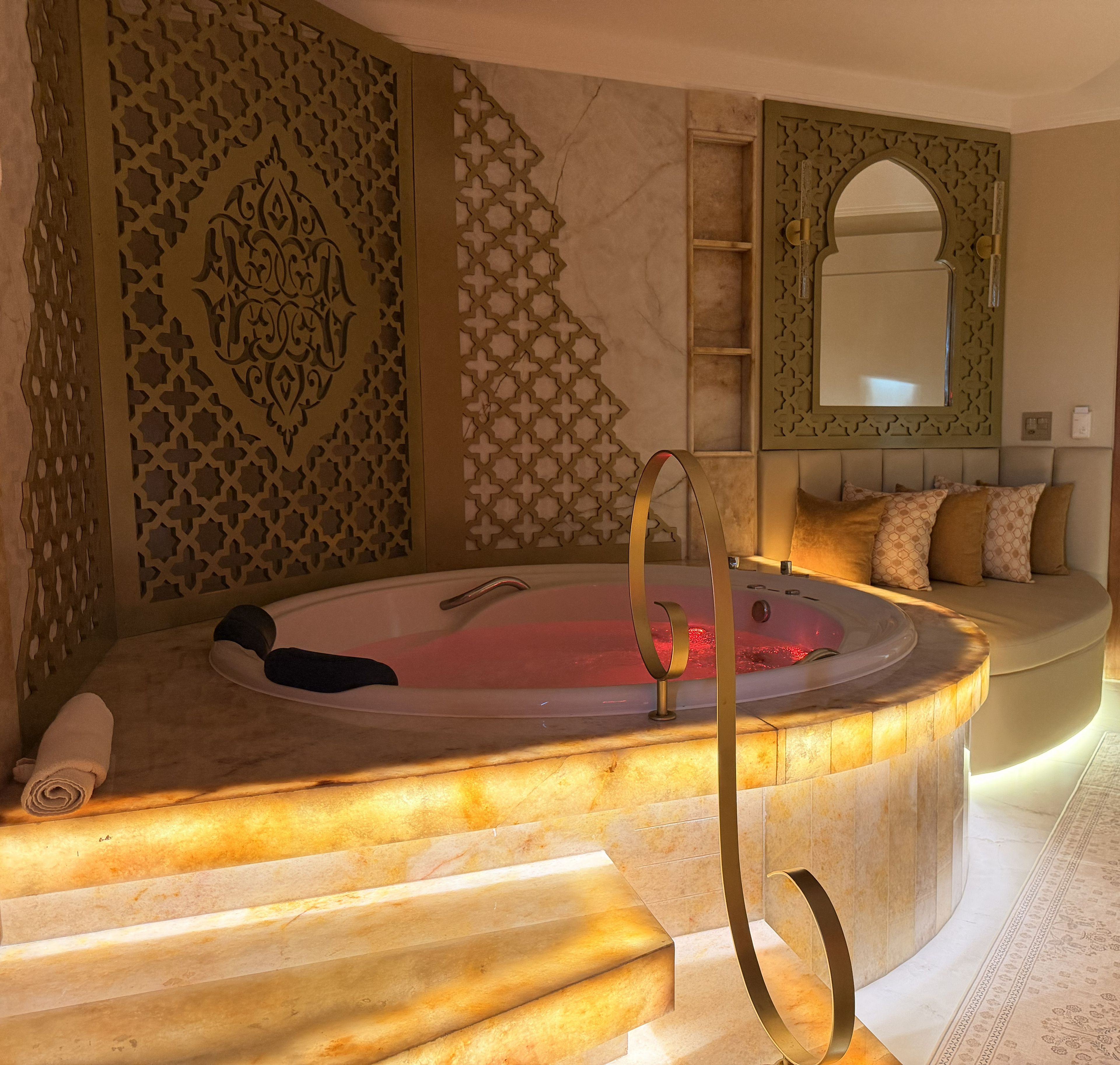 Un cuarto de spa con un jacuzzi, un muro decorativo, una zona de estar con cojines y un espejo.
