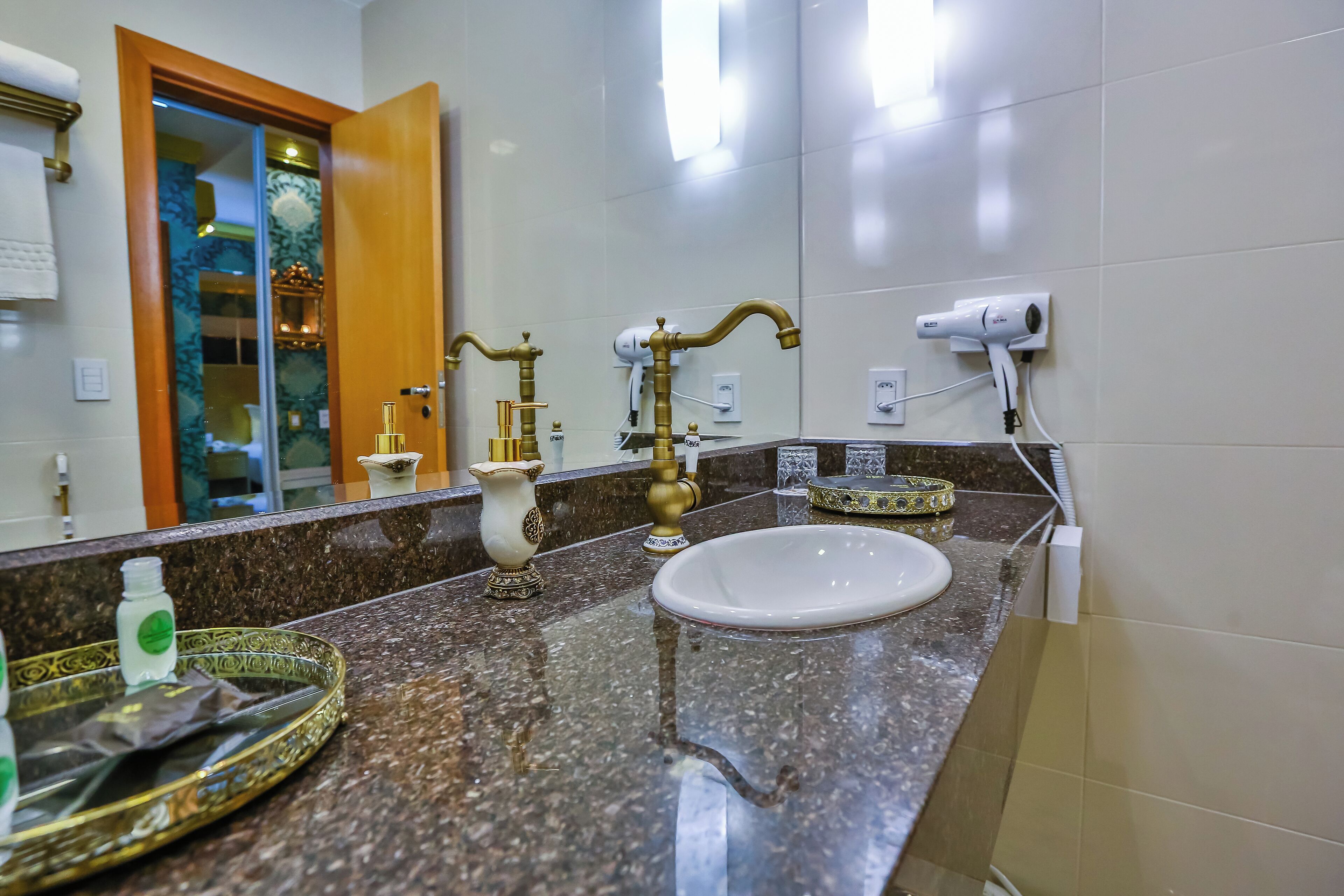 A bathroom with a marble countertop, a white sink, a gold faucet, and a hairdryer mounted on the wall.