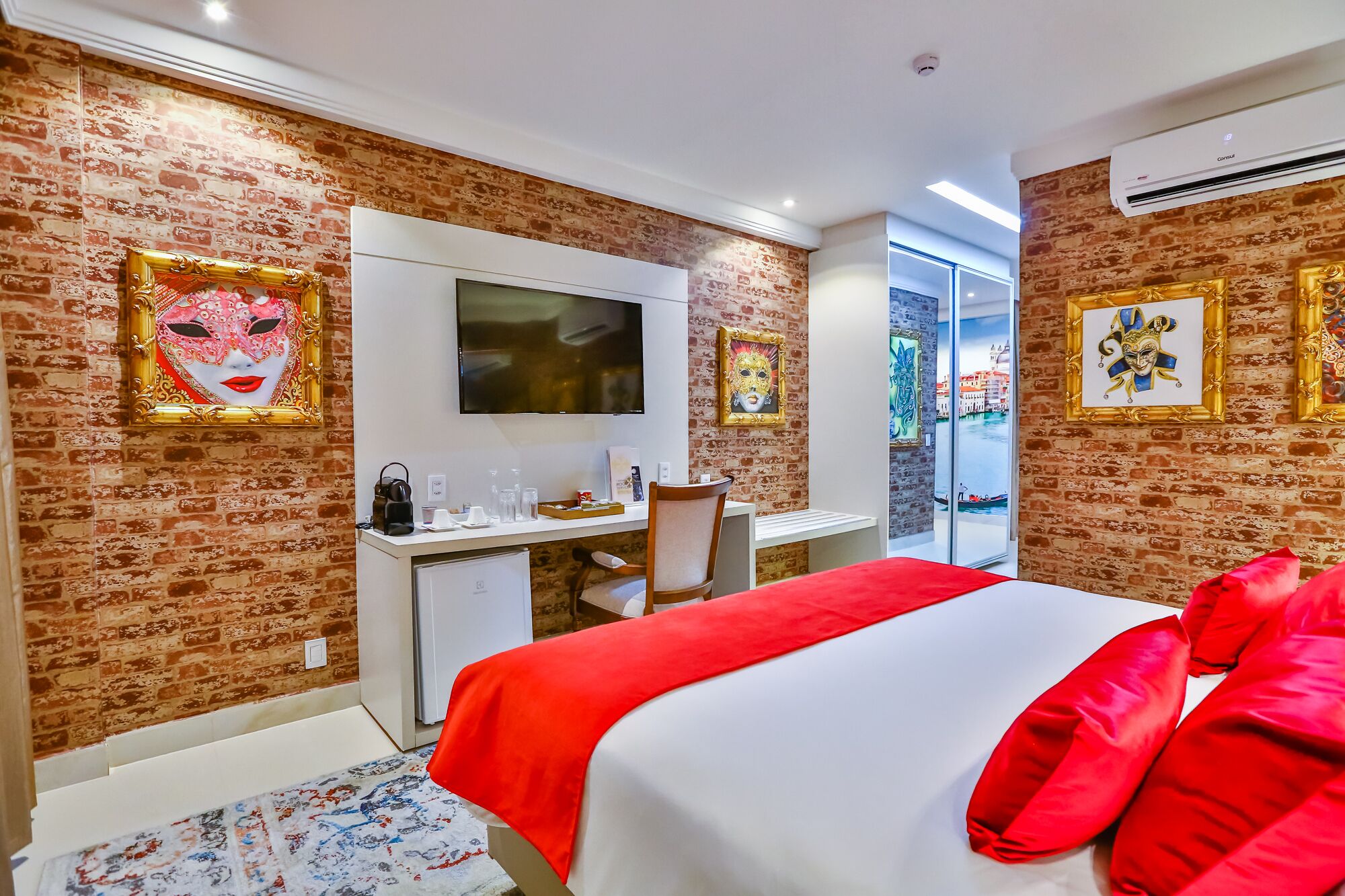 Habitación de hotel con pared de ladrillo, cama con ropa de cama roja, escritorio con computadora, televisor sobre el escritorio y obras de arte en las paredes.