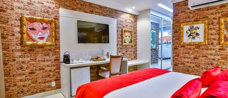 Habitación de hotel con pared de ladrillo, cama con ropa de cama roja, escritorio con computadora, televisor sobre el escritorio y obras de arte en las paredes.
